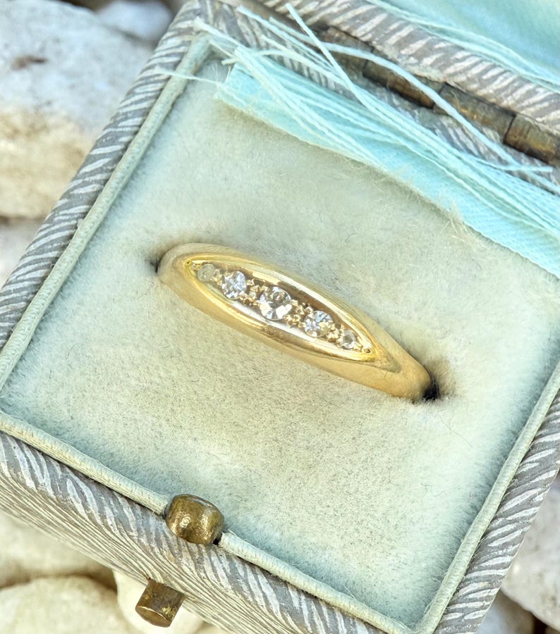 May include: A gold ring with a row of small diamonds set in a curved design. The ring is displayed inside a vintage-style, light blue jewelry box with a decorative clasp and light blue ribbon.