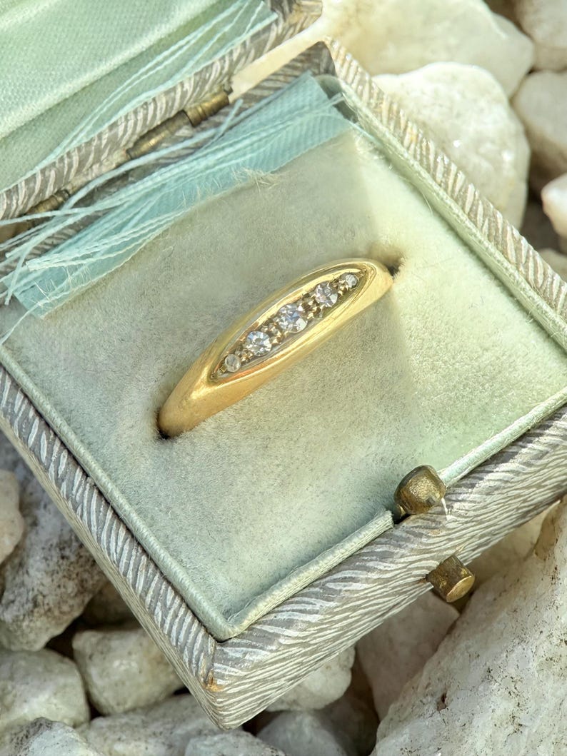May include: A gold ring with a row of small diamonds set in a curved band. The ring is presented in a vintage-style, pale green jewellery box with a patterned exterior and a soft, pale green interior.
