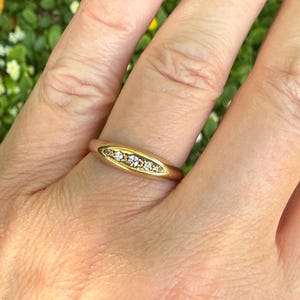 May include: A gold ring with a row of small, clear gemstones set in a horizontal oval shape. The ring is worn on a finger, with the hand in front of a blurred green background.