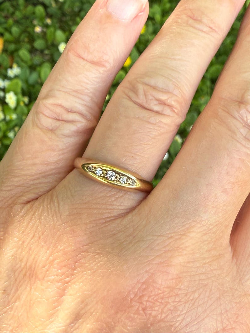 May include: A gold ring with a row of small, clear gemstones set in a horizontal oval shape. The ring is worn on a finger, with the hand in front of a blurred green background.