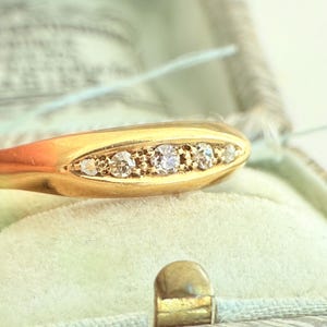 May include: Close-up of a gold ring with five small, clear gemstones set in a row. The ring is displayed inside a vintage, light green jewelry box with a gold clasp. The ring's band is a smooth, polished gold.