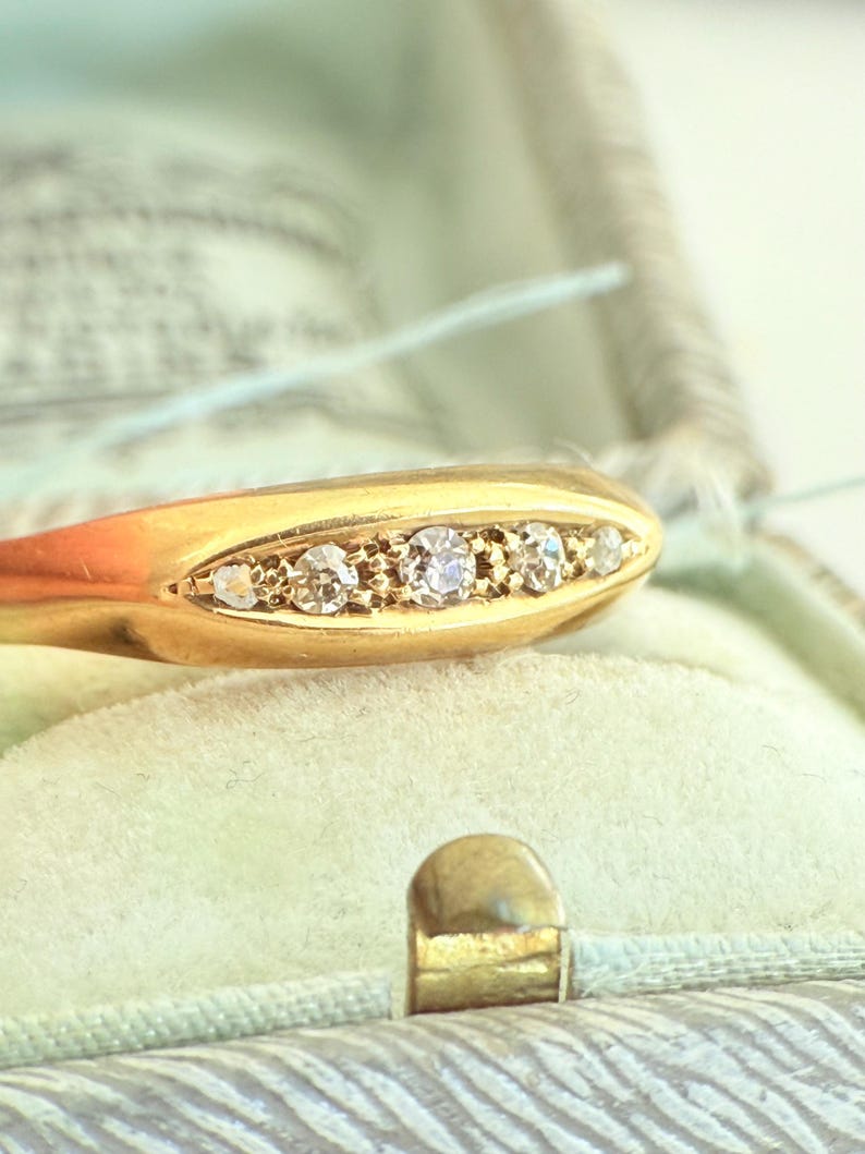 May include: Close-up of a gold ring with five small, clear gemstones set in a row. The ring is displayed inside a vintage, light green jewelry box with a gold clasp. The ring's band is a smooth, polished gold.