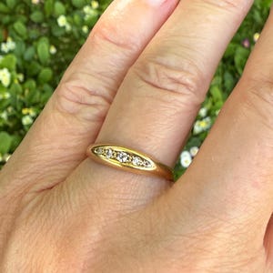 May include: A gold ring with a series of small, clear gemstones set in a row. The ring is on a finger, with a blurred green background of foliage.