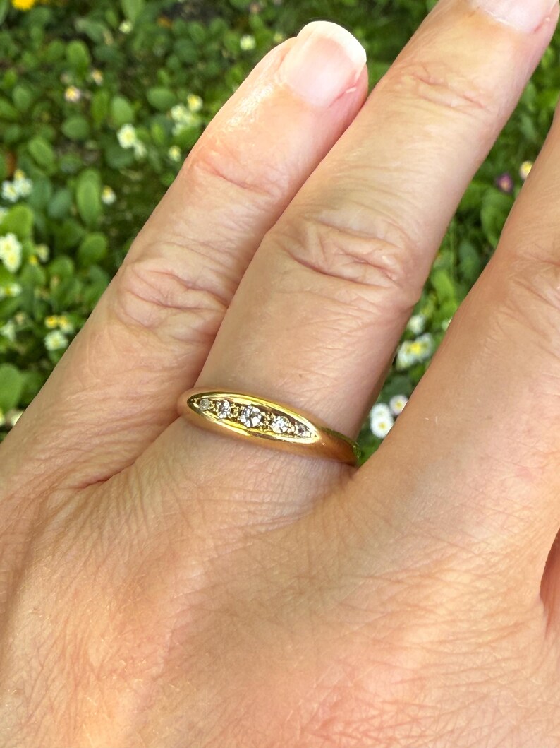 May include: A gold ring with a series of small, clear gemstones set in a row. The ring is on a finger, with a blurred green background of foliage.