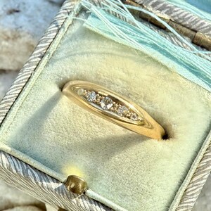 May include: A gold ring with a row of small, clear gemstones set in a curved band. The ring is displayed inside a vintage, light green jewelry box with a decorative lid and a light blue tassel.