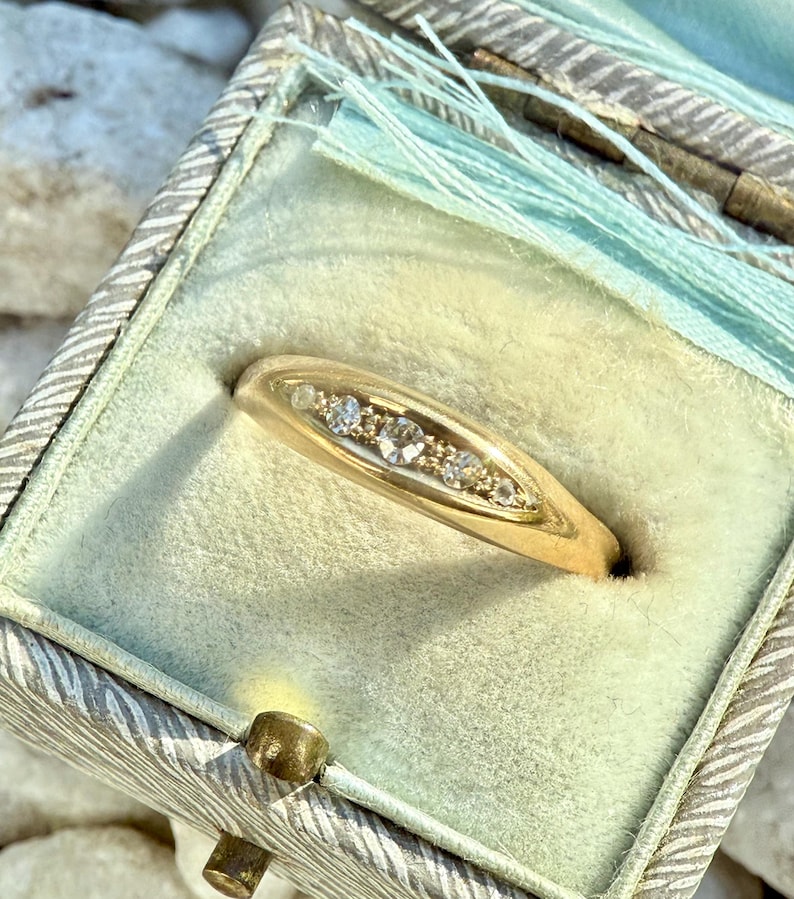 May include: A gold ring with a row of small, clear gemstones set in a curved band. The ring is displayed inside a vintage, light green jewelry box with a decorative lid and a light blue tassel.