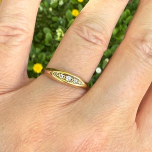 May include: A gold ring with three small, clear gemstones set in a horizontal row. The ring is on a finger, with a blurred green and yellow background.