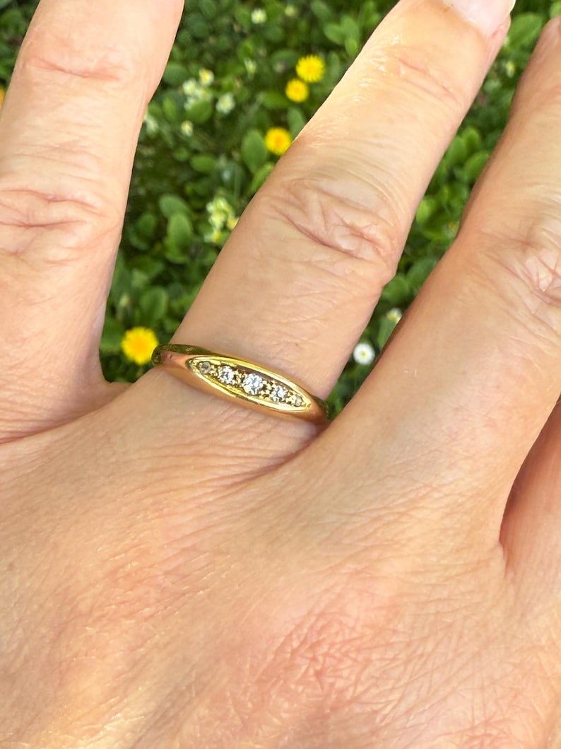 May include: A gold ring with three small, clear gemstones set in a horizontal row. The ring is on a finger, with a blurred green and yellow background.
