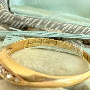 May include: Close-up of a gold ring with small diamonds set along the band. The ring has an inscription that reads "18K". The ring is set against a light blue background.