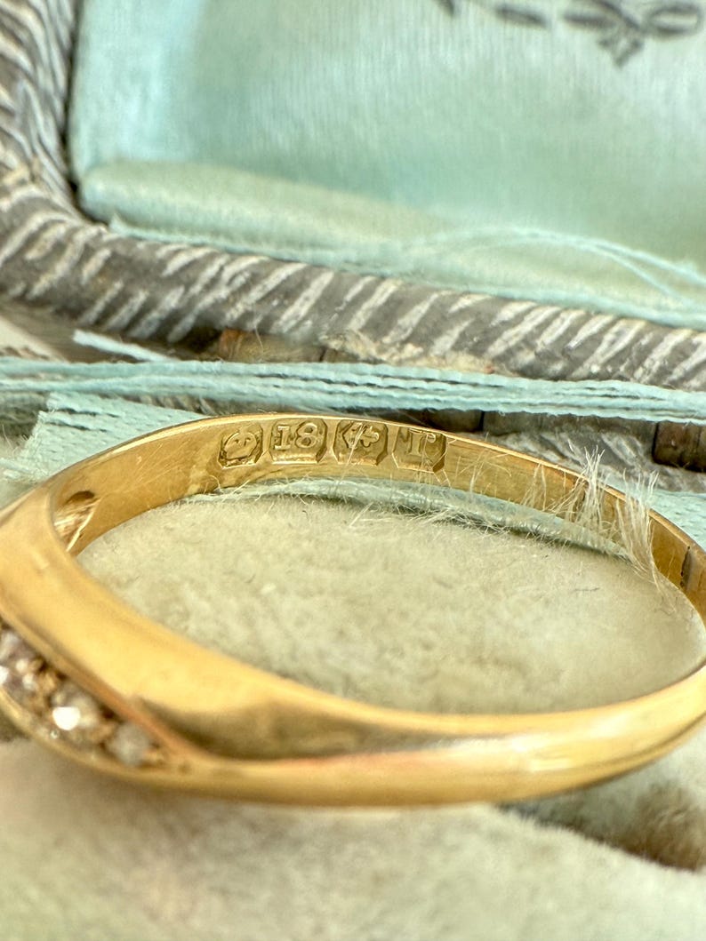 May include: Close-up of a gold ring with small diamonds set along the band. The ring has an inscription that reads "18K". The ring is set against a light blue background.