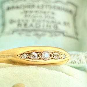 May include: Close-up of a gold ring with five clear gemstones set in a row. The ring is on a light green velvet surface, with a blurred background that includes text. The ring is a piece of jewelry.