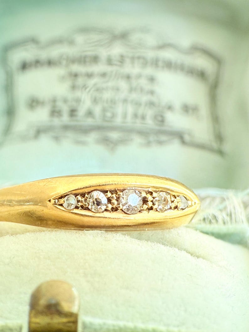 May include: Close-up of a gold ring with five clear gemstones set in a row. The ring is on a light green velvet surface, with a blurred background that includes text. The ring is a piece of jewelry.