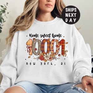 Può includere: Felpa bianca con la scritta "Home sweet home" e i numeri "10001" in un design a tema autunnale. Il design include accenti floreali e di foglie, con le parole "new york, al" sotto.