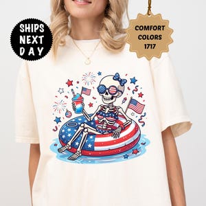 May include: Ivory-colored t-shirt featuring a skeleton in sunglasses and a bow, relaxing on a red, white, and blue inflatable ring. The skeleton is holding a drink and an American flag. The shirt has the text "Ships Next Day" and "Comfort Colors 1717".
