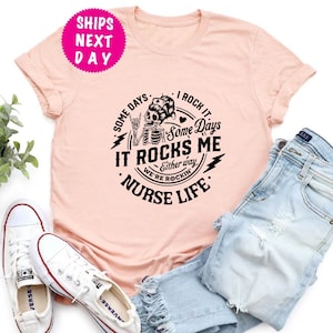 May include: Pink t-shirt with a black graphic design featuring a skeleton wearing a nurse's cap and holding a heart. The text reads "Some Days I Rock It Some Days It Rocks Me Either Way We're Rockin' Nurse Life".