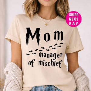 May include: A cream-coloured t-shirt with the words "Mom manager of mischief" in black, along with a lightning bolt and footprints design. The shirt has short sleeves and a crew neck.