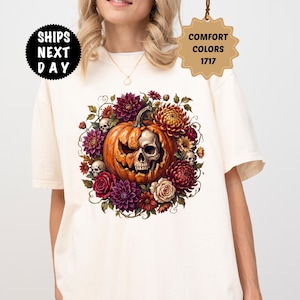 Puede incluir: Una camiseta de manga corta color marfil con un gráfico central de una calabaza tallada con cara de calavera, rodeada por un arreglo circular de dalias coloridas y calaveras pequeñas. La camiseta incluye los textos SHIPS NEXT DAY y COMFORT COLORS 1717.