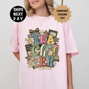 Op de afbeelding: Een lichtroze t-shirt met een grafische print met het woord TEACHER in grote, kleurrijke blokletters. Het ontwerp bevat illustraties van een potlood, een wereldbol, een schoolbus, een beker met het label TEACHER FUEL en sterren met luipaardprint.