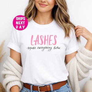 May include: White t-shirt with the word "LASHES" in pink script and the phrase "make everything better" in black script. The shirt is worn with blue denim shorts and a brown belt. A pink circle with "SHIPS NEXT DAY" is in the upper left.