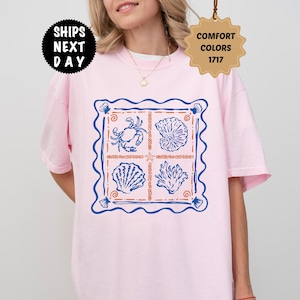 May include: A light pink t-shirt featuring a square graphic print with a blue wavy border. The design contains four sections displaying a blue crab, a seashell, a scallop shell, and coral, separated by orange lines and a small starfish.