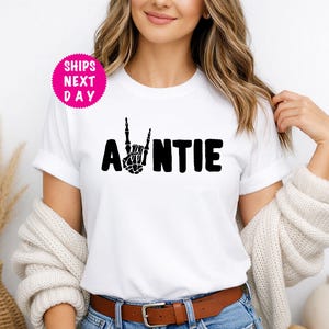Pode incluir: Camiseta branca com a palavra "AUNTIE" em letras pretas em negrito. A letra "N" é substituída por uma mão de esqueleto preta fazendo um sinal de paz. Um círculo rosa com as palavras "SHIPS NEXT DAY" está no canto superior esquerdo.