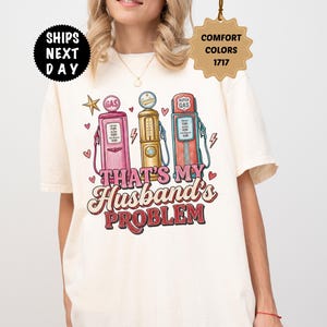 Könnte beinhalten: Elfenbeinfarbenes T-Shirt mit einem Retro-Design von drei Zapfsäulen in Rosa, Gold und Rot. Der Text "THAT'S MY Husband's PROBLEM" ist in einer Vintage-Schriftart mit Herzen und Sternen dargestellt. Das T-Shirt hat einen Rundhalsausschnitt.