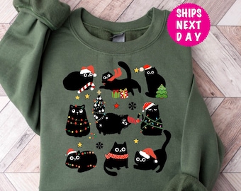 Op de afbeelding: Groen kersttrui met een zwart kattenontwerp. De katten dragen kerstmanmutsen en kerstverlichting. De tekst "Ships Next Day" staat in een roze cirkel in de rechterbovenhoek van de afbeelding.