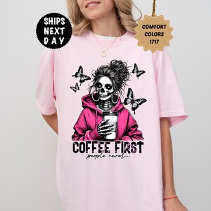 Op de afbeelding: Lichtroze T-shirt met een afbeelding van een schedel met een knot, een roze hoodie draagt, een koffiekopje vasthoudt en omringd is door vlinders. De tekst "COFFEE FIRST people never..." staat eronder.