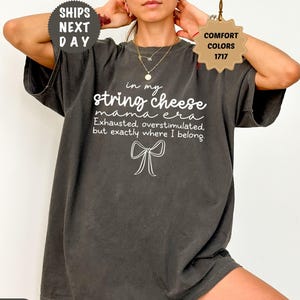 May include: Pepper-colored t-shirt with the text "in my string cheese mama era" in white. The shirt also features the phrases "Exhausted, overstimulated, but exactly where I belong" and a bow graphic. The shirt has a relaxed fit.