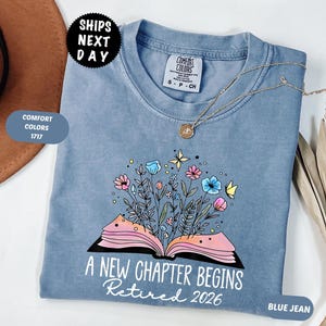 May include: A blue jean Comfort Colors t-shirt with the text "A New Chapter Begins Retired 2026." The shirt features a floral design with an open book. A gold necklace is draped across the shirt.