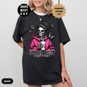 Op de afbeelding: Zwart T-shirt met een skeletafbeelding met een roze hoodie, met een koffiekopje. Het ontwerp bevat vlinders en de tekst "COFFEE FIRST people never..." Het shirt heeft een ronde hals en korte mouwen.