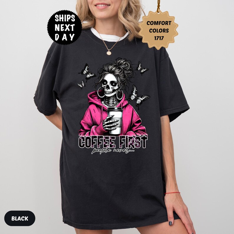 Op de afbeelding: Zwart T-shirt met een skeletafbeelding met een roze hoodie, met een koffiekopje. Het ontwerp bevat vlinders en de tekst "COFFEE FIRST people never..." Het shirt heeft een ronde hals en korte mouwen.