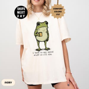 Puede incluir: Una camiseta de manga corta color marfil con una ilustración gráfica de una rana verde sosteniendo una taza amarilla. Debajo de la rana, el texto dice: "i have to say weird stuff or i'll die." La camiseta tiene cuello redondo y un corte holgado.
