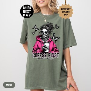 Op de afbeelding: Mosgroen T-shirt met een afbeelding van een schedel met een roze hoodie en een koffiekopje. De tekst "COFFEE FIRST people never..." staat eronder. Vlinders omringen de schedel. Het shirt heeft een relaxte pasvorm.