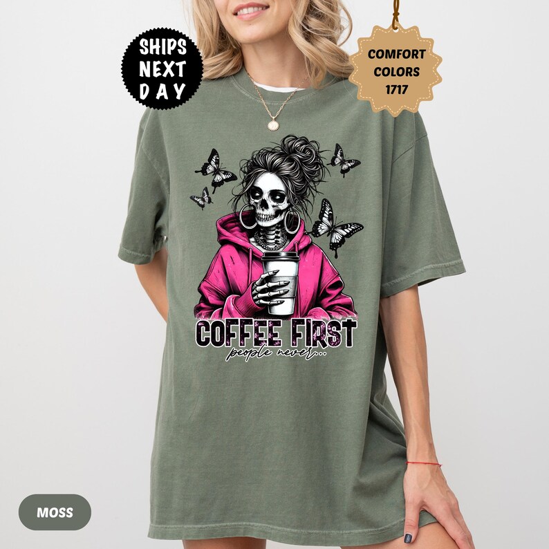 Op de afbeelding: Mosgroen T-shirt met een afbeelding van een schedel met een roze hoodie en een koffiekopje. De tekst "COFFEE FIRST people never..." staat eronder. Vlinders omringen de schedel. Het shirt heeft een relaxte pasvorm.