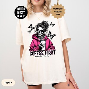 Op de afbeelding: Ivoor kleurig T-shirt met een afbeelding van een skelet met een roze hoodie en een koffiekopje, met vlinders. De tekst "COFFEE FIRST people never..." staat eronder. Het shirt heeft korte mouwen.