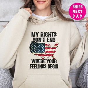 Op de afbeelding: Een beige hoodie met een verweerde Amerikaanse vlag afbeelding en de tekst "Mijn rechten eindigen niet waar jouw gevoelens beginnen".