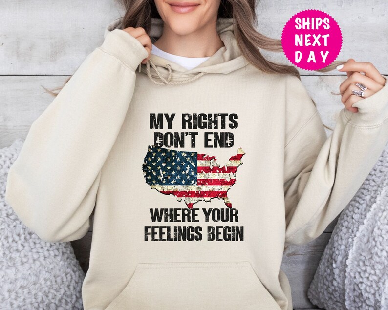 Op de afbeelding: Een beige hoodie met een verweerde Amerikaanse vlag afbeelding en de tekst "Mijn rechten eindigen niet waar jouw gevoelens beginnen".