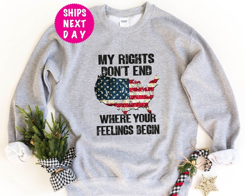 Op de afbeelding: Grijs sweatshirt met een verweerd Amerikaans vlagontwerp en de tekst "Mijn rechten eindigen niet waar jouw gevoelens beginnen".