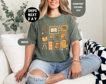 May include: A moss green short-sleeve t-shirt featuring a graphic print with various autumn-themed illustrations including an open book, a pumpkin, a mug, a slice of pie, and a stack of books, all arranged in a grid with orange and yellow accents.