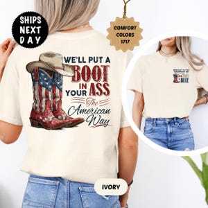 May include: Ivory-colored t-shirt featuring a graphic of cowboy boots with an American flag design and a cowboy hat. The text reads "WE'LL PUT A BOOT IN YOUR ASS The American Way." Additional text includes "SHIPS NEXT DAY" and "COMFORT COLORS 1717."