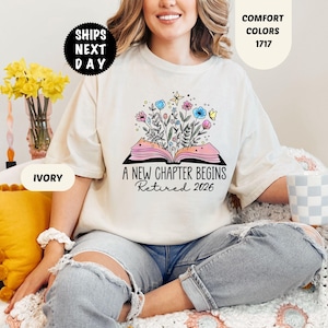 May include: Ivory-colored t-shirt with the text "A New Chapter Begins Retired 2026" and a floral design over an open book. The shirt is a comfort color and is being worn by a person.