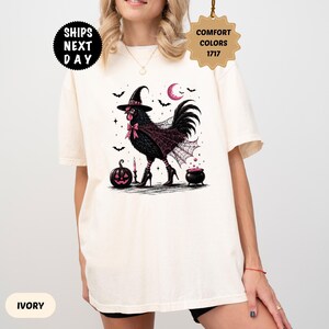 May include: An ivory t-shirt featuring a graphic of a black chicken wearing a witch hat, a spiderweb cape, and high-heeled boots. The design includes a pink crescent moon, bats, a jack-o'-lantern, a candle, and a small cauldron with pink bubbles.