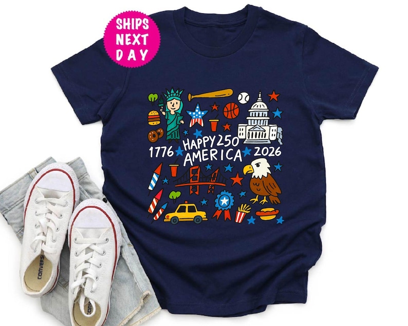 Puede incluir: Camiseta azul marino con un gr&aacute;fico colorido que celebra el 250 aniversario de Am&eacute;rica. El dise&ntilde;o incluye la Estatua de la Libertad, un bate de b&eacute;isbol, una pelota de baloncesto, el Capitolio y el texto "Happy 250 America 2026".