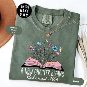 May include: A moss green t-shirt with the text "A New Chapter Begins Retired 2026" and a floral book design. The shirt has a crew neck and short sleeves. A gold necklace is draped on the shirt.