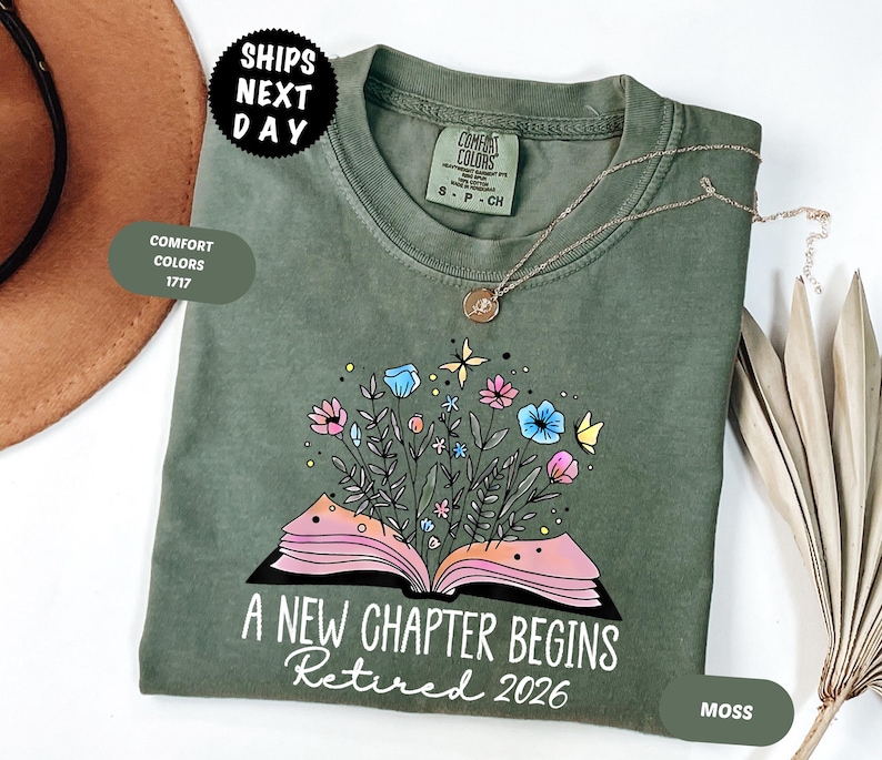 May include: A moss green t-shirt with the text "A New Chapter Begins Retired 2026" and a floral book design. The shirt has a crew neck and short sleeves. A gold necklace is draped on the shirt.