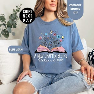 May include: Blue jean colored t-shirt with the text "A New Chapter Begins Retired 2026". The shirt features a graphic of an open book with flowers and butterflies. The shirt is a comfort color.