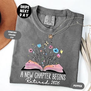 May include: A gray t-shirt with a floral book design. The design features an open book with colorful flowers and butterflies. The text "A New Chapter Begins Retired 2026" is printed below the book.
