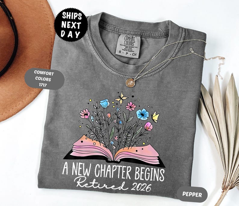 May include: A gray t-shirt with a floral book design. The design features an open book with colorful flowers and butterflies. The text "A New Chapter Begins Retired 2026" is printed below the book.