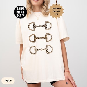 May include: An ivory short-sleeve t-shirt featuring a graphic print of three horse bit snaffles arranged vertically. The top bit has a black and white checkerboard pattern, the middle has a leopard print, and the bottom has a black polka dot pattern.